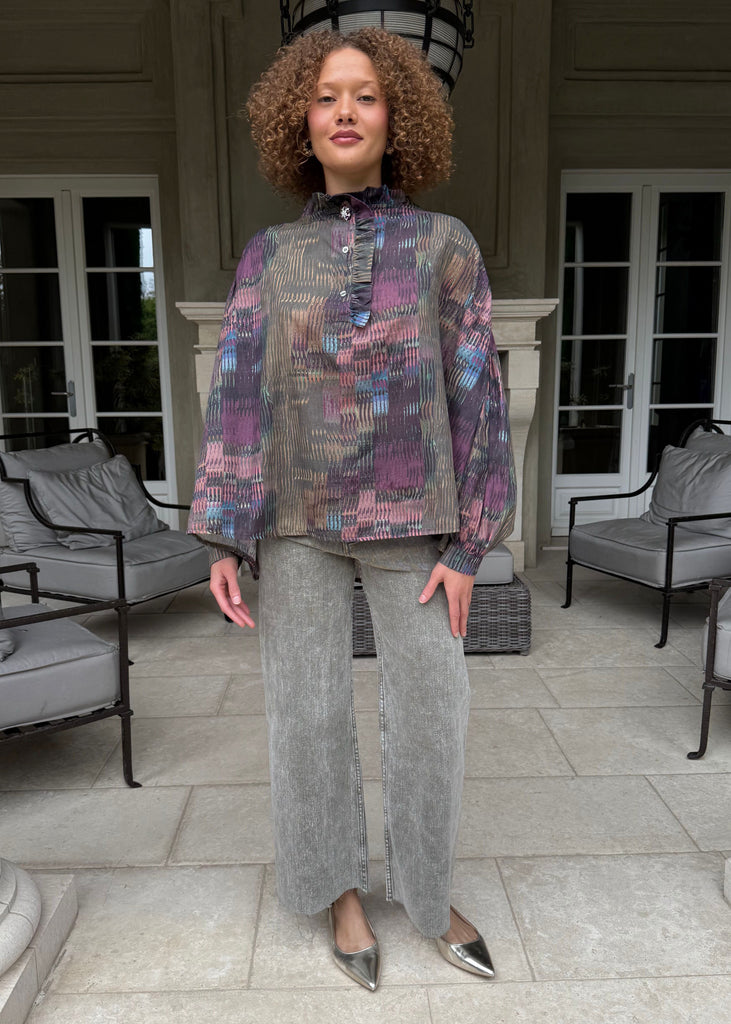 Woman standing in a stylish outfit with a patterned top and gray pants on a patio.