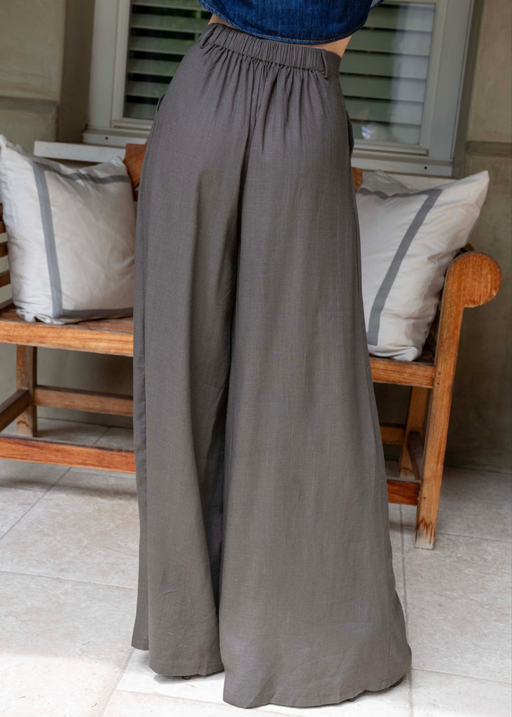 Person wearing gray wide-leg pants standing in a room with wooden furniture and cushions.