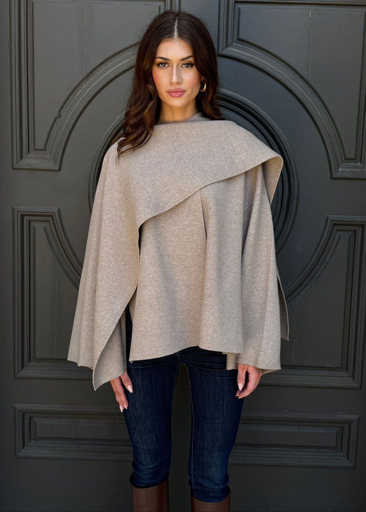 Woman wearing a beige poncho and dark jeans standing in front of a gray door.