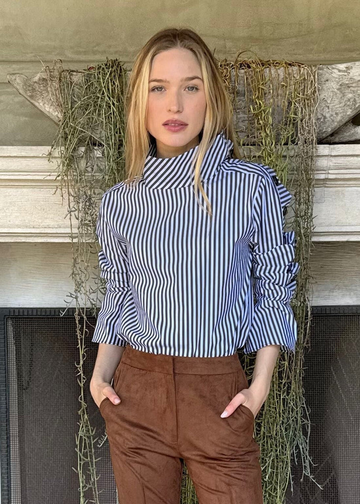 Woman standing in front of a fireplace wearing a blue striped shirt and brown pants.