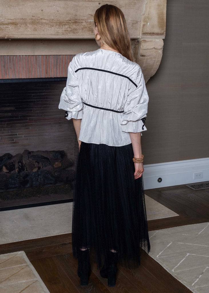 Woman standing in front of a fireplace wearing a white blouse with black stripes and a black skirt.